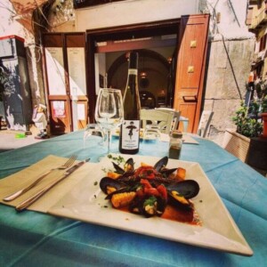 All'aperto davanti all'Antica Osteria Pisano, tavolo con tovaglia azzurra, piatto rettangolare di pesce e cozze, bottiglia di vino bianco e bicchieri, menu visibile all'ingresso.