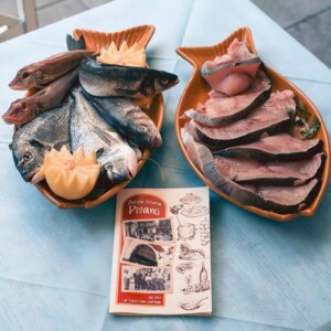 Pesci interi e fette di pesce disposti in vassoi arancioni, decorati con scorze di limone, su tavolo azzurro; brochure Antica Osteria Pisano visibile al centro.
