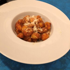 Gnocco dorato in ciotola bianca con spolverata di parmigiano, servito su tavolo azzurro; richiamo alla cucina napoletana dell’Antica Osteria Pisano.