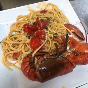 Spaghettoni con astice e pomodorini, parmigiano grattugiato, prezzemolo e scorza di limone su piatto bianco; carne d'astice rossa brillante e pasta lunga.