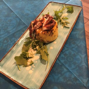 Tortino al centro del piatto decorato con tentacolo di polpo grigliato, salsa cremosa e foglie di misticanza su un piatto azzurro allungato.