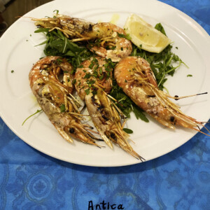 Gamberi cotti al forno o alla griglia serviti su foglie di rucola con spicchi di limone e prezzemolo, in piatto bianco, contesto di tavolo colorato; rimando alla cucina di Antica Osteria Pisano
